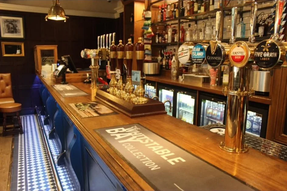 Finished pub bar with a polished timber bar top, brass beer fonts and a blue panelled bar front lit above a chequered tiled floor strip.