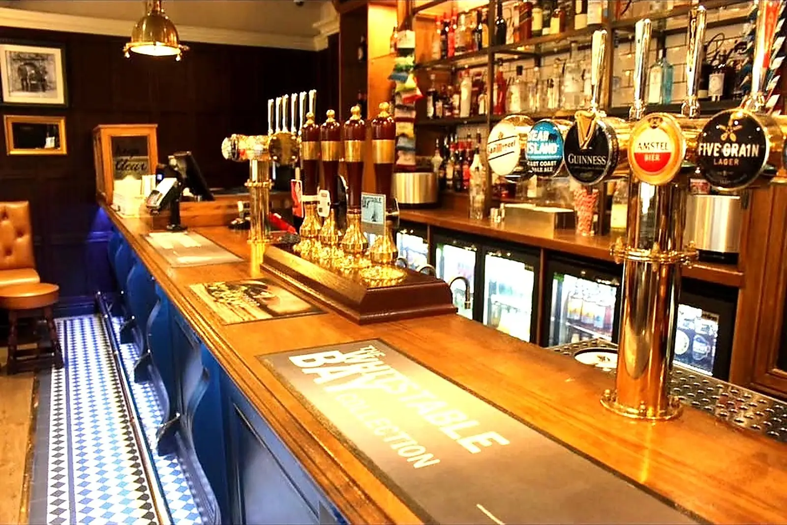 Finished pub bar with a polished timber bar top, brass beer fonts and a blue panelled bar front lit above a chequered tiled floor strip.