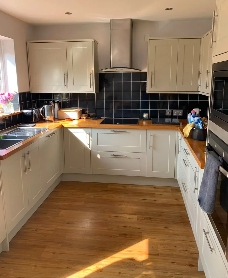 A cream shaker kitchen fitted in a U shape with oak worktops, a black tiled splashback and a chimney extractor over the hob.
