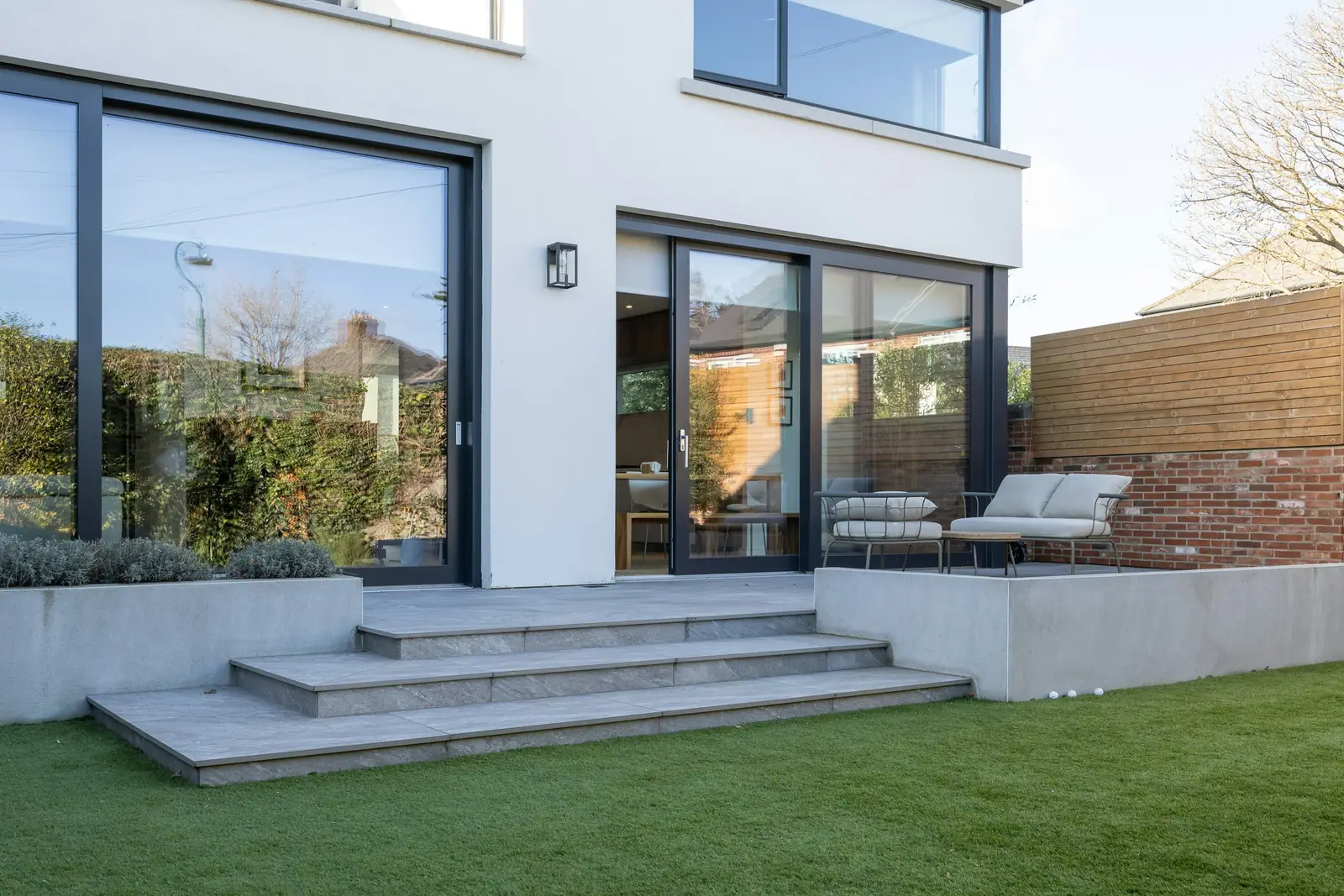 A white-rendered rear extension with full-height dark sliding doors onto a paved terrace and steps down to a lawn.