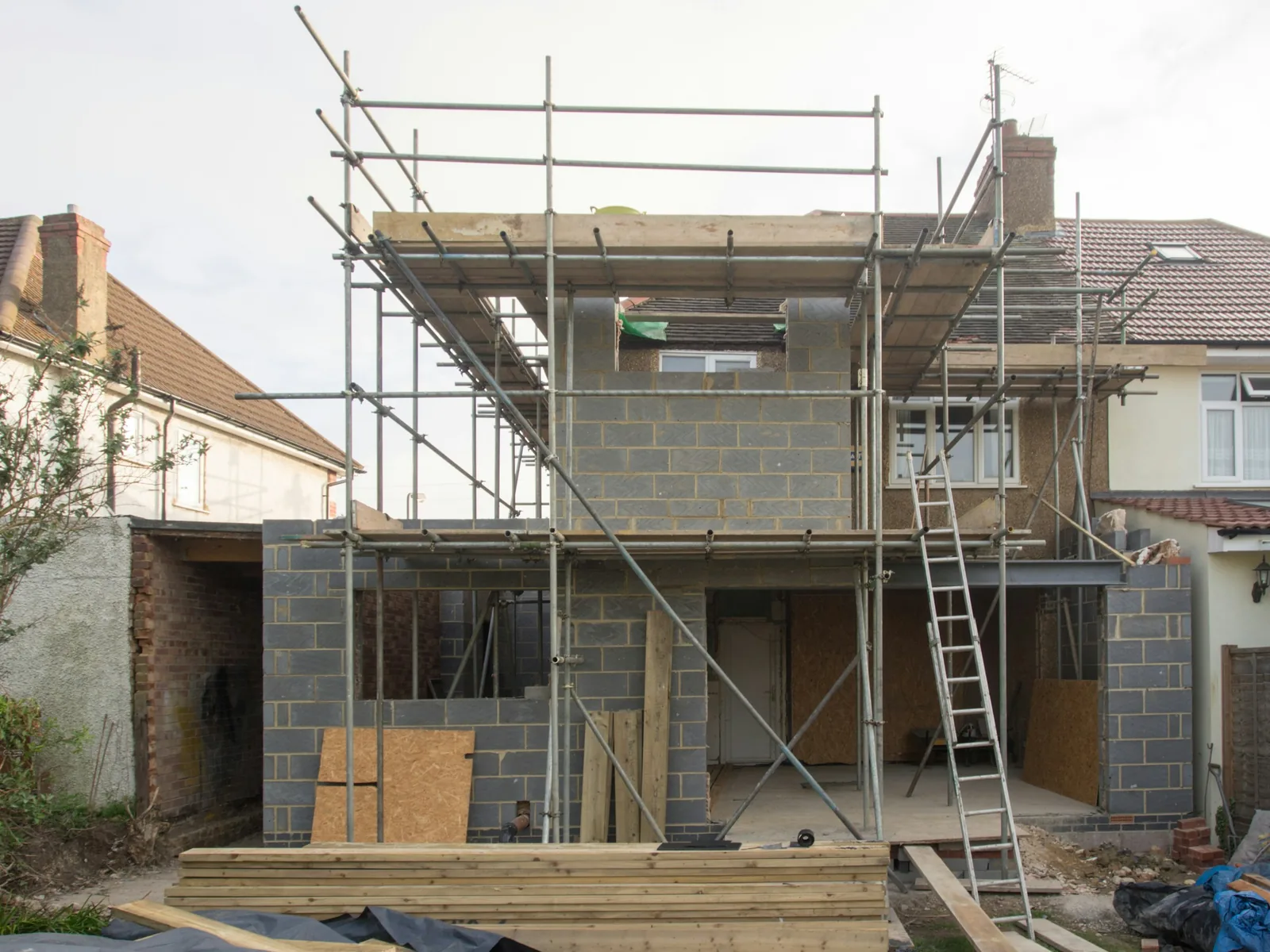 A two-storey rear extension under construction on a pebbledash semi, its blockwork shell wrapped in scaffolding.