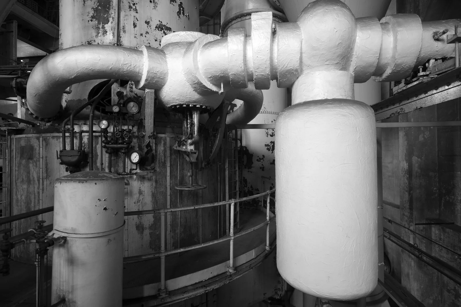 Lagged pipework, gauges and handwheel valves above a railed walkway in an old plant room.