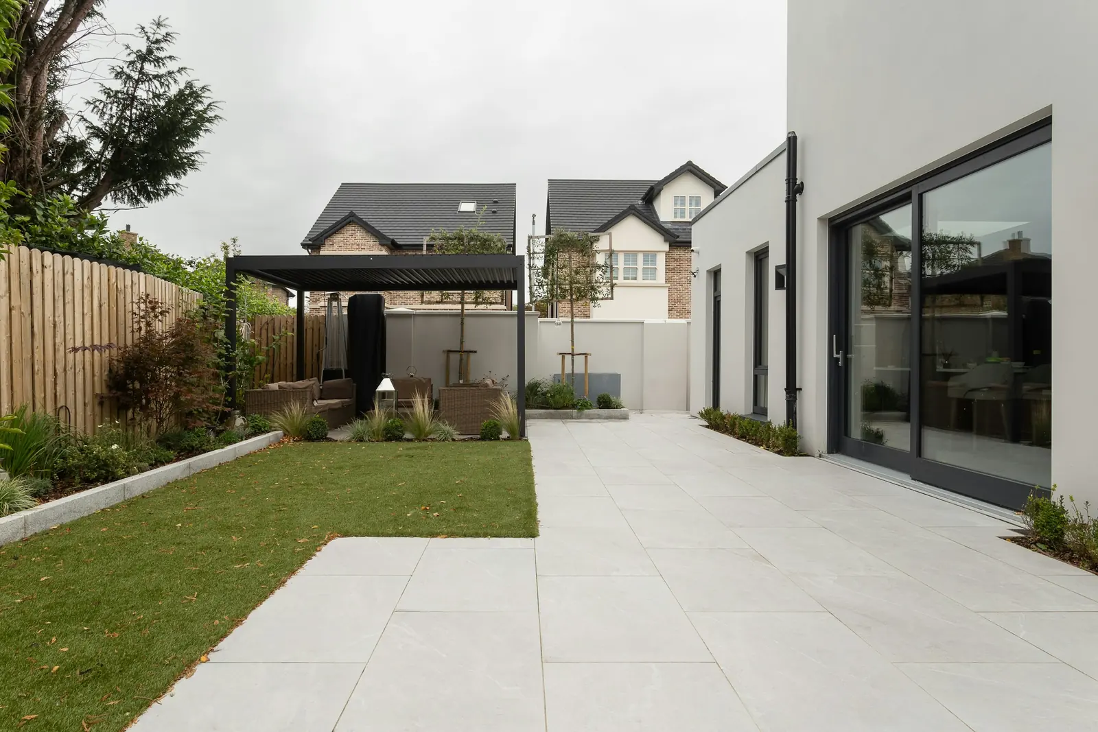 Dark-framed sliding doors in a rendered rear extension, pale slab paving running out to the lawn.